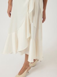 Λευκή μίντι φούστα με ασύμμετρο στρίφωμα, φορεμένη με μπεζ ψηλές σαγιονάρες σε ανοιχτό φόντο.