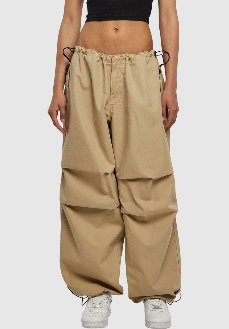 Personne portant un pantalon beige à jambes larges avec cordon à la taille et aux chevilles, un crop top noir et des baskets blanches sur un fond uni.