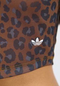Brun gennemsigtig top med sort og grå leopardprint. Har gule syninger og et hvidt broderet logo nær kanten.