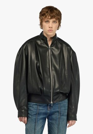 Persona con capelli corti e castani che indossa una giacca in pelle nera oversize con cerniera e jeans blu su uno sfondo semplice.