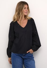 Marine-blaues Oberteil mit V-Ausschnitt, verfügt über einen einfarbigen Oberkörper und schwarze Lochmuster-Puffärmel. Trägt es mit Jeans, präsentiert in einem neutralen Umfeld.