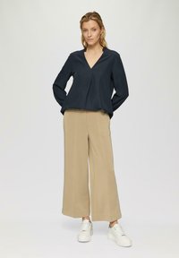Top de manga larga azul marino con escote en V y pliegue en la parte delantera, combinado con pantalones beige de pierna ancha y zapatillas blancas. Tejido suave con un corte relajado.