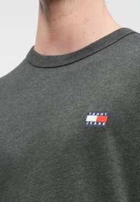 Grå bomull T-skjorte med rund hals og korte ermer. Har et brodert "TOMMY JEANS"-logo i blått, hvitt og rødt på brystet.