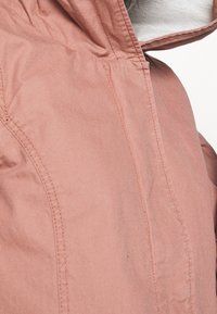 Růžová lehká bunda s kapucí, hladkou texturou, viditelným šitím a uvolněným střihem. Reflexní detail blízko zipu.