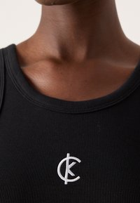 Svart ribbad linne med rund halsringning, med en vit broderad logotyp på bröstet. Slät textur med figursydd design.