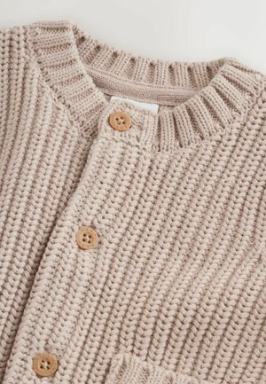 Vaaleanbeige neulottu neuletakki, jossa on resorireunainen pääntie, puiset napit ja teksturoitu kirjailu. Mukana on myös paikkatasku.