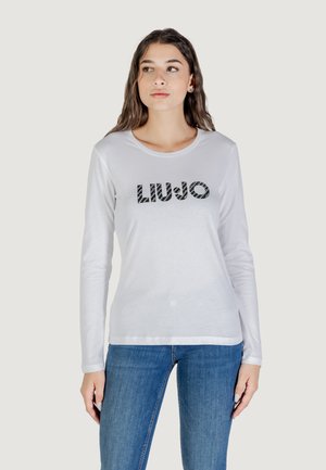 Il modello indossa una camicia a maniche lunghe di colore grigio chiaro con la scritta "LIU.JO" in un design audace, abbinata a jeans blu, su uno sfondo neutro.