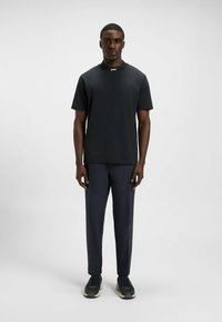 Man staat rechtop met het gezicht naar voren, gekleed in een zwart BOSS shirt met korte mouwen, marineblauwe broek en zwarte sneakers, tegen een eenvoudige witte achtergrond.