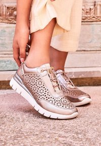 Zapatillas metálicas en color oro rosa con paneles de malla perforada, suelas blancas, diseño de cordones y un acabado liso en la puntera y el talón.
