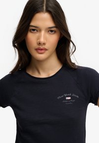 Joven con el pelo largo y castaño, lleva una camiseta negra con el texto "Departamento de Atletismo del Estado de Nueva York" y una pequeña bandera estadounidense en el pecho.