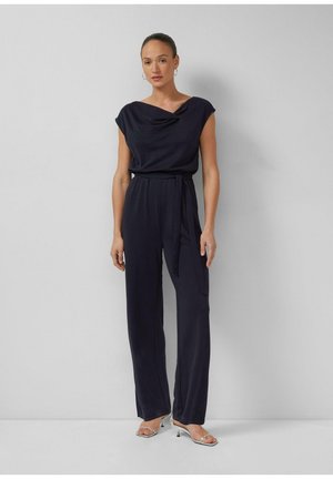 Kobieta w granatowym kombinezonie bez rękawów z kołnierzem w formie fałd oraz paskiem w talii, w srebrnych sandałach na obcasie, stojąca na tle jasnoszarej ściany.