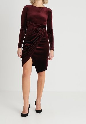 Kobieta w dopasowanej, bordowej welwetowej sukience z długim rękawem, z marszczonym detalem i asymetrycznym dołem, w połączeniu z czarnymi szpilkami.