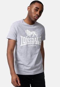 Tricou din bumbac gri, cu un logo alb de leu și textul "Lonsdale London". Mâneci scurte, decolteu rotund și o croială lejeră.