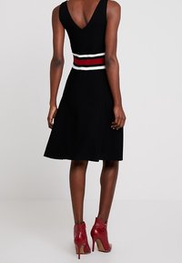 Robe noire à encolure en V et jupe évasée, avec une ceinture rayée contrastante en blanc et rouge. Portée avec des bottines rouges.