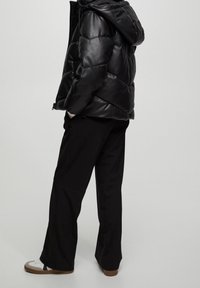 Chaqueta de plumas negra con capucha, con un diseño acolchado y dobladillo elástico. Se lleva con pantalones negros ajustados y zapatos con suela marrón.