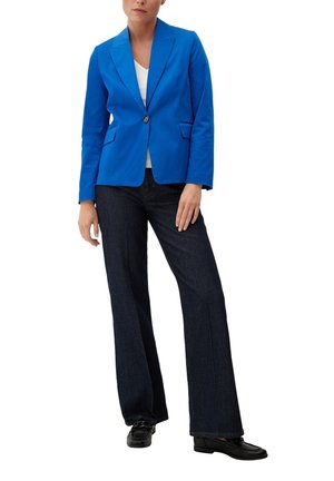 Vrouw draagt een felblauwe getailleerde blazer, wit topje, donkere wijde jeans en zwarte loafers, staand met één hand ontspannen langs haar zij.