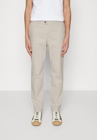 Person som bär beige raka byxor, vit t-shirt och vita sneakers med gröna accenter, ståendes mot en enfärgad bakgrund.