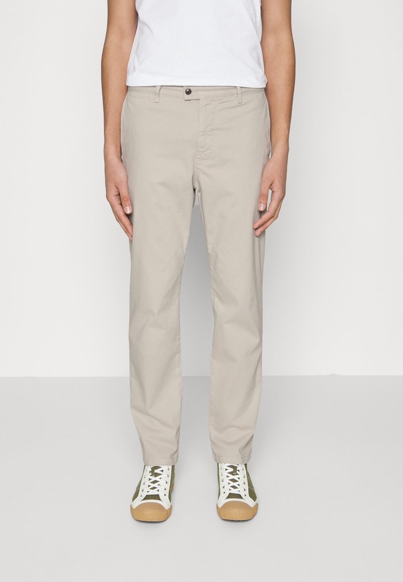 Person som bär beige raka byxor, vit t-shirt och vita sneakers med gröna accenter, ståendes mot en enfärgad bakgrund.