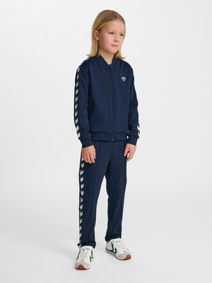 Barnet står iført en mørkeblå træningsdragt med hvide chevronstriber på ærmer og ben, hvide sneakers med grønne accenter.