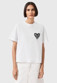 Hvid bomulds t-shirt med korte ærmer, rund halsudskæring og en sort hjerte grafik på venstre bryst. Blød tekstur, afslappet pasform.
