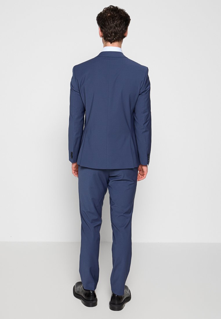 Abito blu navy con una vestibilità su misura, caratterizzato da un solo spacco e tessuto liscio. I pantaloni sono a taglio dritto con tasche piatte, indossati con scarpe nere.