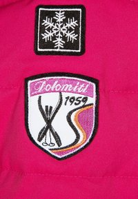Il tessuto fucsia presenta due toppe ricamate: una fiocco di neve nero e un design stilizzato con sci incrociati, "Dolomiti", e l'anno "1959".