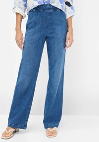 Wide-leg denim trousers in medium blue. Features a smooth texture, waistband with loops, and subtle stitching details. Paired with light sandals.