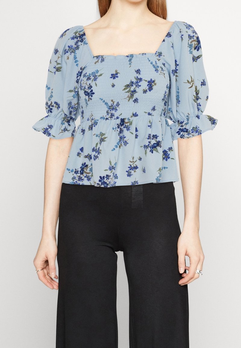 Femme portant un chemisier floral bleu clair avec des manches ballon et un corsage froncé, associé à un pantalon noir à jambes larges.