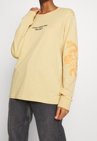 Langärmliges, hellgelbes Baumwoll-T-Shirt mit einem schwarzen Textdesign auf der Brust und einer orangefarbenen Schlange sowie einem Blütenmotiv auf dem linken Ärmel.