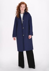 Marineblauer langer Mantel aus glattem Stoff, mit einem spitzen Kragen, silbernen Knöpfen und hochgerollten Ärmeln. Getragen über schwarzen Hosen.