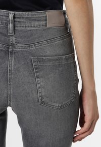 Jean en denim gris au tissu lisse, avec une poche arrière et un petit patch en cuir de la marque sur la ceinture. Coupe ajustée.