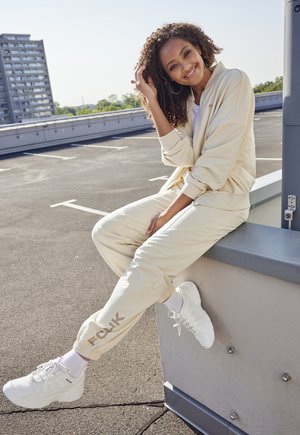Cream-colored tracksuit with a zip-up jacket and elastic pants, paired with white sneakers. Features FCK logo on pants, smooth fabric, relaxed fit.