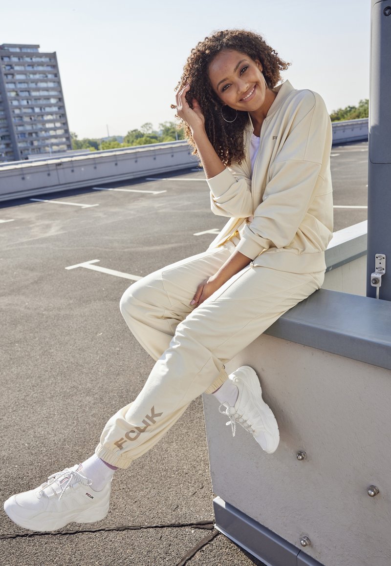 Crème-kleurig trainingspak met een ritsjack en elastische broek, gecombineerd met witte sneakers. Bevat het FCK-logo op de broek, van gladde stof en met een relaxed fit.