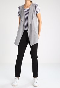 Šedá bezrukávová vesta s volným designem přes černo-bílé pruhované tričko, spárovaná s černými kalhotami a bílými teniskami.