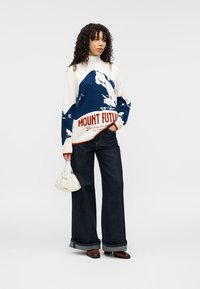 Pull en tricot oversize crème avec un motif de montagne bleu et le texte rouge "MOUNT FUTURE". Associé à un jean large foncé et un sac blanc.