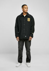 Jachetă din fleece texturat, neagră, cu front cu nasturi, guler și un mare logo a literei "S" de culoare aurie. Combinată cu blugi închiși la culoare și adidași albi.