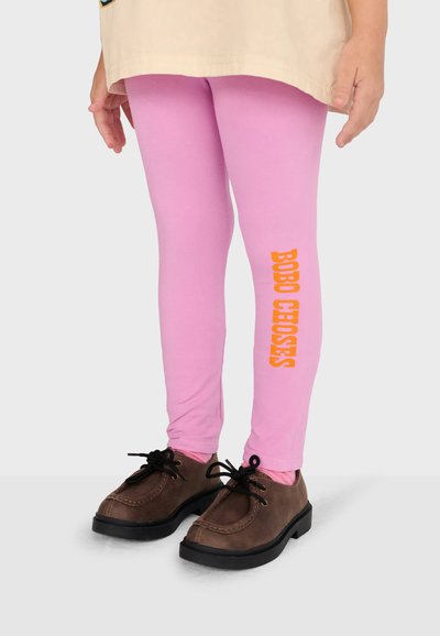 Bambino che indossa leggings rosa chiaro con testo arancione "BOBO CHOSES" sulla gamba e scarpe marroni con lacci, in piedi con le mani rilassate lungo i fianchi.