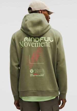 Oliven grøn hættetrøje med et stort "Mindful Movement" grafisk motiv i lysegrønne og røde mønstre på ryggen. Blød tekstur, afslappet design og løs pasform.