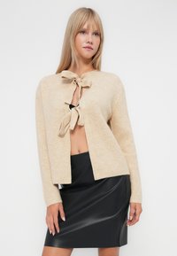 Bežs adīts kardigāns ar saitēm, atvērts priekšpuse, ribota tekstūra un garas piedurknes, apvienots ar melnu pieguļošu ādas svārku.