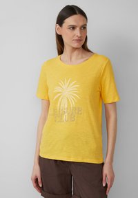 T-shirt jaune à manches courtes en tissu doux, avec un motif de palmier et le texte "LEISURE CLUB" en gris clair. Encolure ronde.