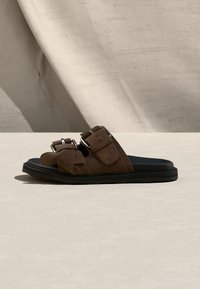 Καφέ σανδάλι με σαγιονάρα με δύο ρυθμιζόμενες λουρίδες με αγκράφες και μαύρη σόλα σε ανοιχτό ουδέτερο φόντο.