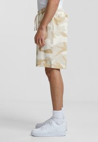 Beige camo shorts with an elastic waistband and drawstring. Made of soft fabric, featuring a side pocket and a straight cut. Paired with white sneakers.