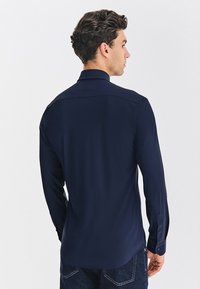 Granatowa koszula z długim rękawem, z kołnierzykiem i mankietami zapinanymi na guziki. Charakteryzuje się gładką fakturą materiału i lekko zaokrąglonym dołem.