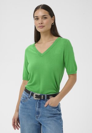 Donna che indossa una maglietta verde brillante a maniche corte con scollo a V infilata nei jeans blu con una cintura nera, in posa con una mano in tasca.