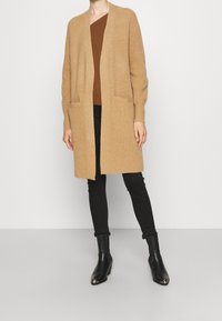 Beige stickad cardigan med öppen front, långa ärmar och två fickor, lager på lager över en brun topp och svarta skinnybyxor, kombinerad med svarta ankelstövlar.