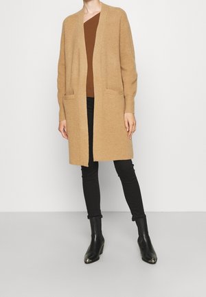 Cárdigan de punto beige con apertura frontal, mangas largas y dos bolsillos, superpuesto sobre una blusa marrón y pantalones ajustados negros, complementado con botas negras de tobillo.