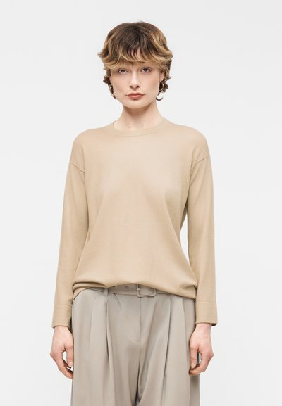 Personne aux cheveux courts et ondulés portant un pull beige à manches longues rentré dans un pantalon plissé gris clair taille haute avec une ceinture.