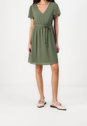 Femme portant une robe kaki longueur genou avec des manches volantes et une ceinture nouée à la taille, assortie à des chaussures plates beiges à boucles.