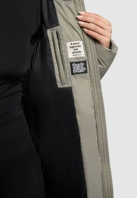 Mano che tiene aperto un giacca beige con fodera nera, mostrando due etichette in tessuto con il testo del marchio vicino alla tasca interna.
