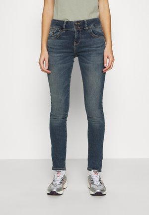 Skinny jeans in dark blue denim, featuring a mid-rise waist, two front pockets, and orange stitching. Paired with gray athletic shoes.